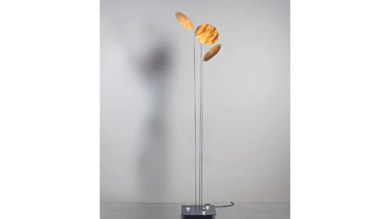 Flavia Thumshirn assina para Ingo Maurer a luminária de piso Nuunu, com estrutura minimalista e cúpula em papel metalizado na cor dourada.