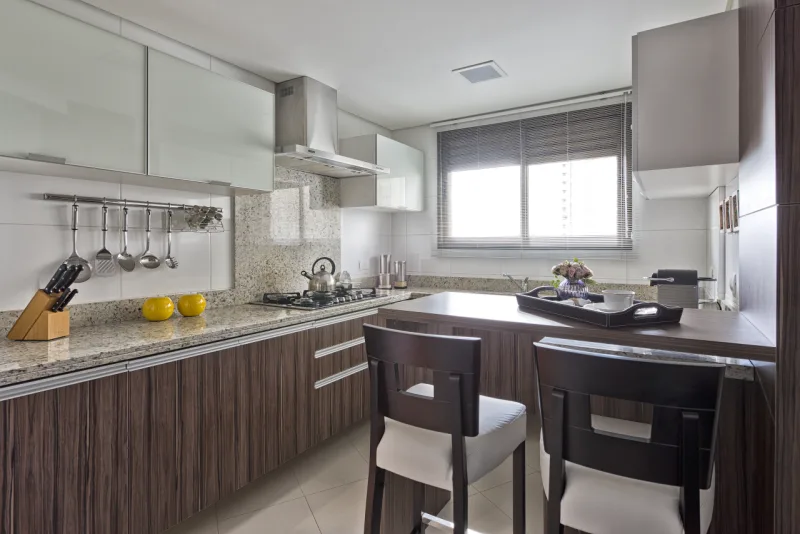 Na cozinha, em continuidade a sala de jantar, foi utilizado o tom madeirado escuro nos armários e branco criando contraste com os eletrodomésticos. Uma bancada central foi criada para descontrair na hora do preparo e facilitar o dia a dia.