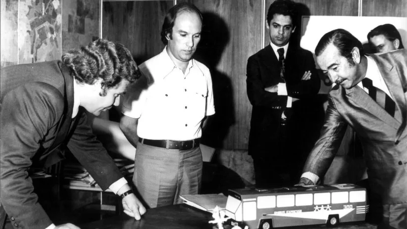 Prefeito Jaime Lerner (esq.), Hairton Romani (centro) e um diretor da Marcopolo, durante apresentação do protótipo do novo ônibus Expresso, na sala de reunião do IPPUC. O modelo foi projetado em conjunto pelo setor de planejamento e a encarroçadora de Caxias do Sul. Os veículos começaram a circular no eixo Norte/Sul, ligando Santa Cândida ao Capão Raso, a partir de 22 de setembro de 1974.